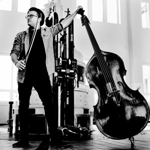 Adam Ben Ezra standing inside the hydroelectric power plant holding his double bass and fiddlestick.