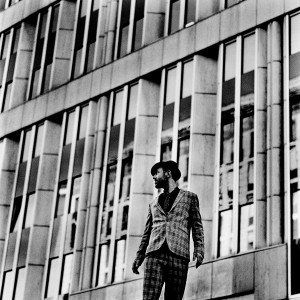 Charlie Winston standing in front of an office building looking back wearing hat and a suit.