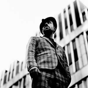 Charlie Winston standing in front of an office building wearing a hat and a suit.