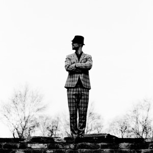 Charlie Winston standing on old brick wall with trees in the background wearing suit and a hat.