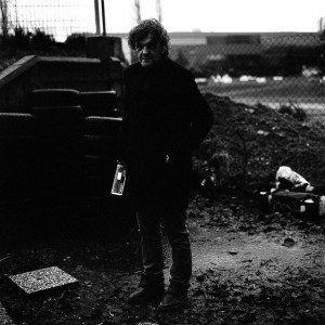Emir Kusturica standing wearing black and holding a bottle of alcohol.