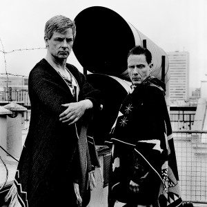 Gus Gus members Daniel Agust Haraldsson and Birgir Porarinsson posing at two tubes on a rooftop of a building with city view in the background.