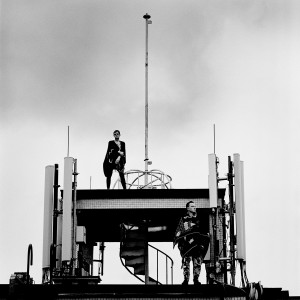 Gus Gus members Daniel Agust Haraldsson and Birgir Porarinsson standing on a broadcaster at the rooftop of the building with skies in the background.