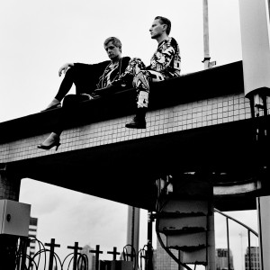 Gus Gus members Daniel Agust Haraldsson and Birgir Porarinsson sitting on a rooftop floor above the stairs wearing high heels.