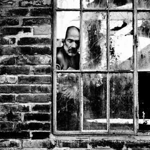 Jean-Marc Barr standing behind an old broken window in a brick house.