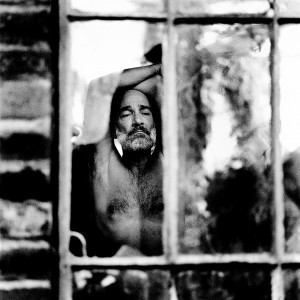 Naked Jean-Marc Barr making a pose with his hands up behind an old broken window in a brick house.