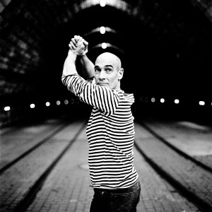 Jean-Marc Barr posing and making gesture in front of a tunnel wearing striped t-shirt.