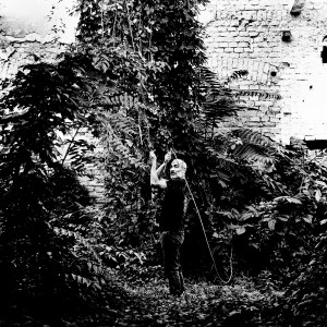 Jean-Marc Barr posing with a giant plants in a jungle house wearing black.