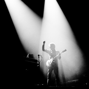 Jean-Michel Jarre playing guitar on the stage at his concert with one hand up.