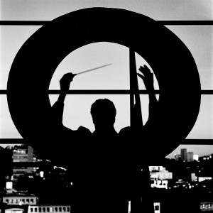 Kevin Griffiths in a conducting pose with his hands up holding baton on the rooftop of a building with giant letter O and the city view in the background.