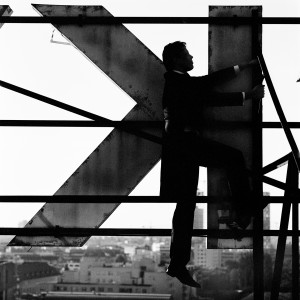 Kevin Griffiths climbing on a giant letter K at the rooftop of a building with city view in the background.