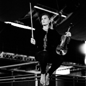 Mari Samuelsen sitting barefoot on the railing with violin and fiddlestick in her hands.