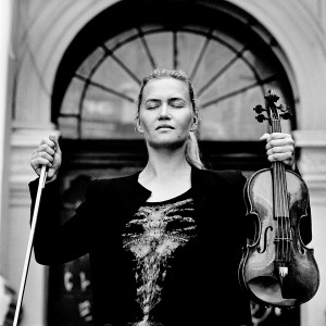 Mari Samuelsen with eyes closed in front of a door holding violin and a fiddlestick.