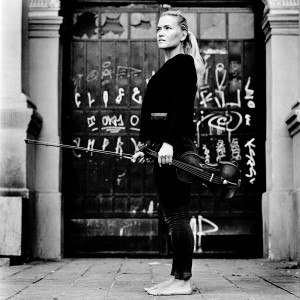 Mari Samuelsen standing barefoot in front of door with graffiti holding violin and fiddlestick.  