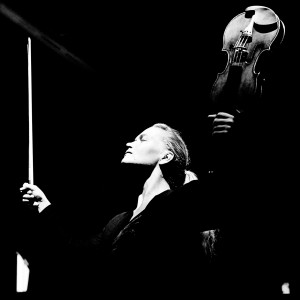 Mari Samuelsen making a gesture with her hands up holding a violin and a fiddlestick in the darkness.