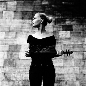 Mari Samuelsen holding violin looking to the left with brick wall in the background.