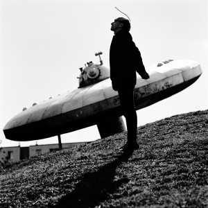 Cyborg artist Neil Harbisson standing at statue of ufo spaceship.