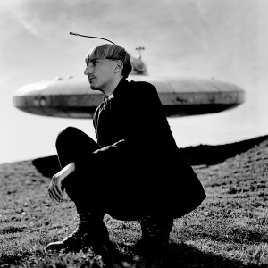 Cyborg artist Neil Harbisson squating in front of ufo spaceship monument.