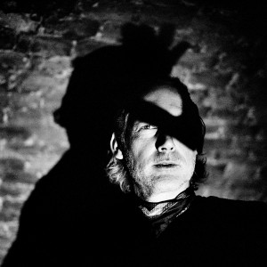 Face of Richard Dorfmeister with one eye covered with the shadow of his hand with the brick wall in the background.